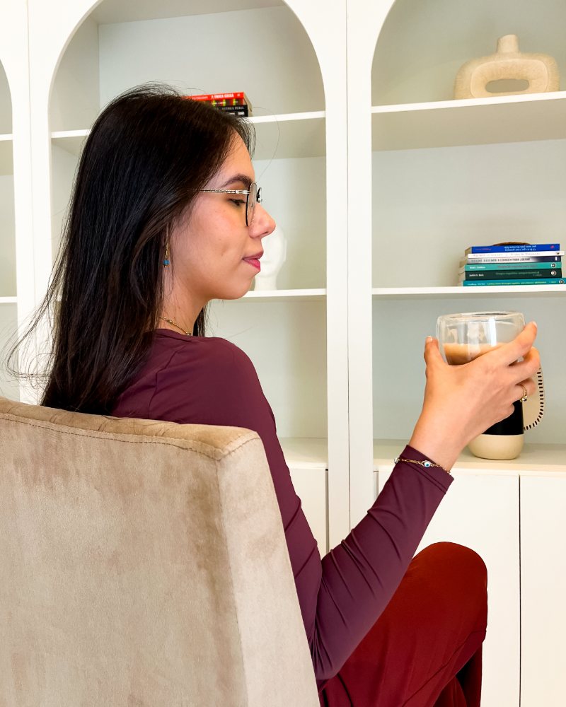 Fernanda Manuela, psicóloga, segurando uma xícara de café em frente à estante do consultório